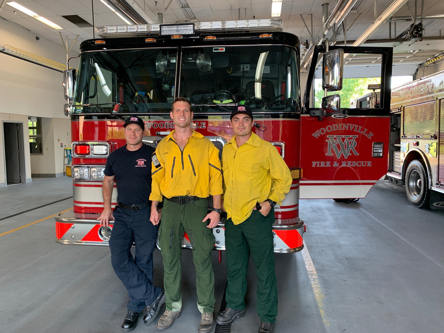 Three firefighters standing in front of a fire truck in a garage wearing Krosen's Uniforms